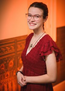 A young woman wearing glasses and a deep red dress smiles warmly while posing indoors against a softly lit orange background. She stands with her hands gently clasped, exuding confidence and joy after participating in the Poetry Out Loud competition.