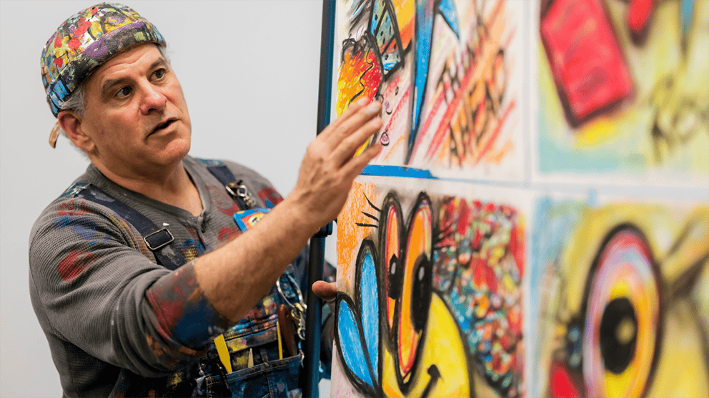 An artist wearing a colorful hat and shirt showing off his paintings