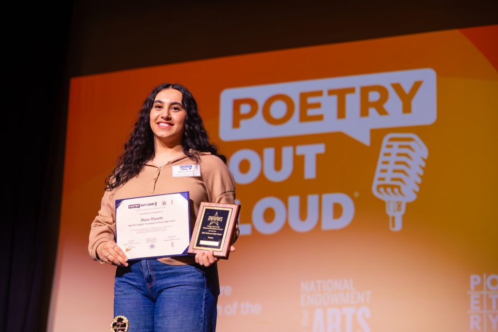 A smiling Poetry Out Loud participant stands onstage holding a certificate and a plaque, with the large “Poetry Out Loud” logo and microphone graphic projected on an orange backdrop behind them.