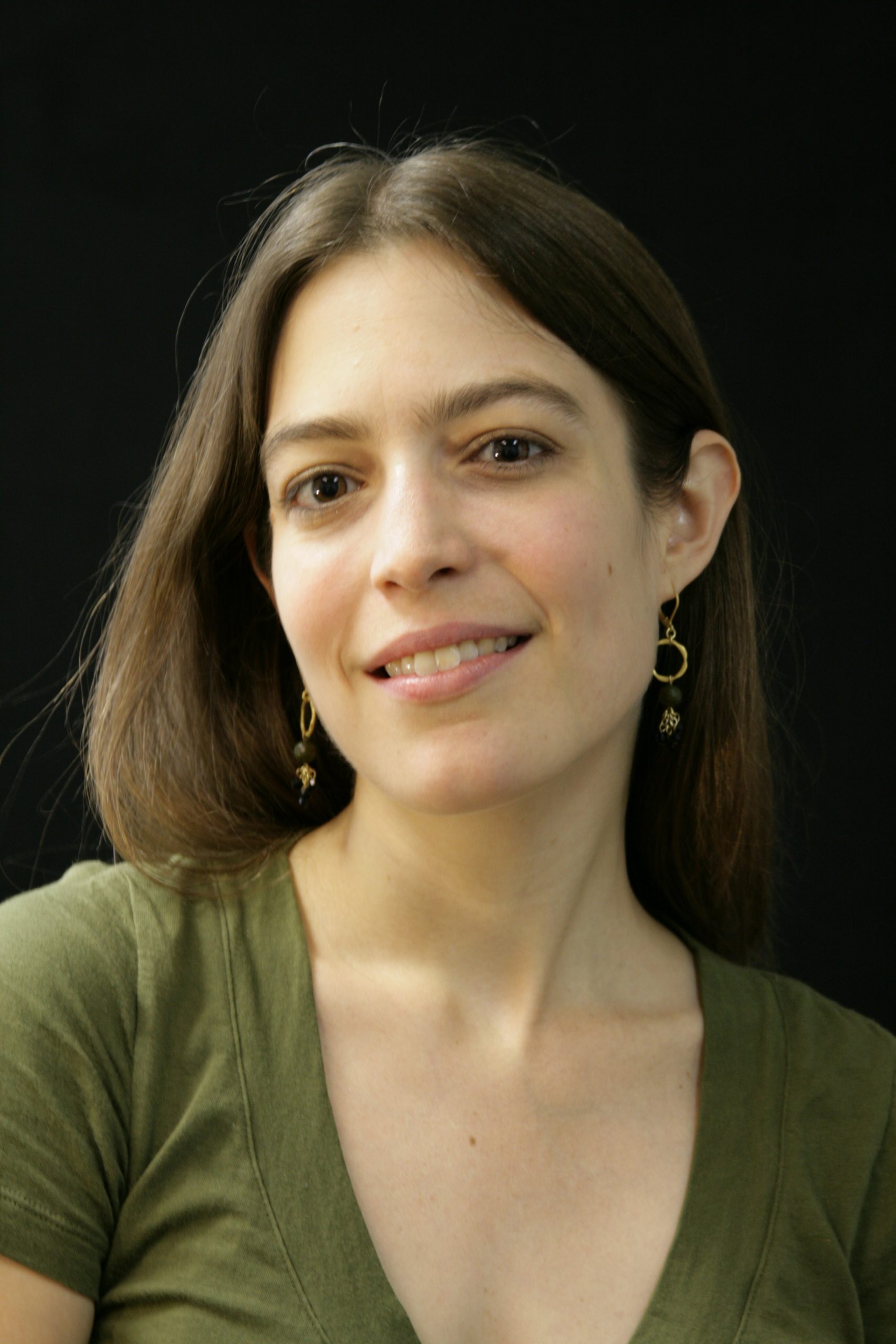 Close-up portrait of a woman with long dark brown hair, wearing a green top and delicate dangling earrings. She faces the camera with a soft, relaxed expression against a simple dark background.