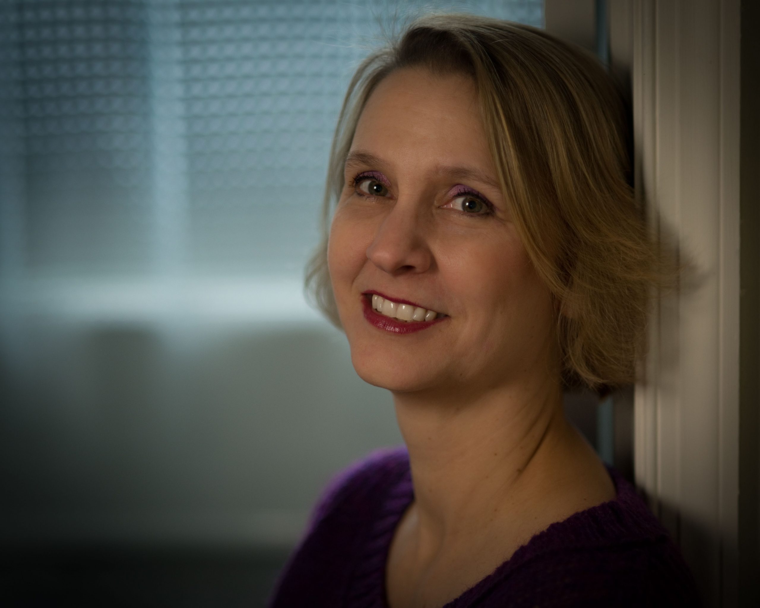 Close-up portrait of a smiling woman with short blonde hair, leaning against a wall or doorframe. Soft light illuminates her face, and the blurred background gives the image a warm, calm, and intimate feel.