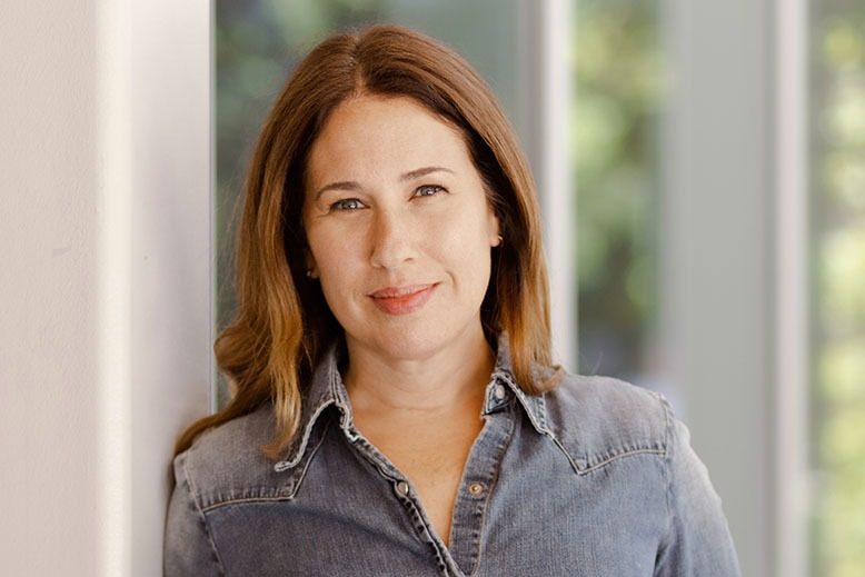 Portrait of a woman with long brown hair leaning against a light-colored wall, wearing a denim button-up shirt. She looks directly at the camera with a soft smile, while bright windows and blurred greenery create a clean, natural background.
