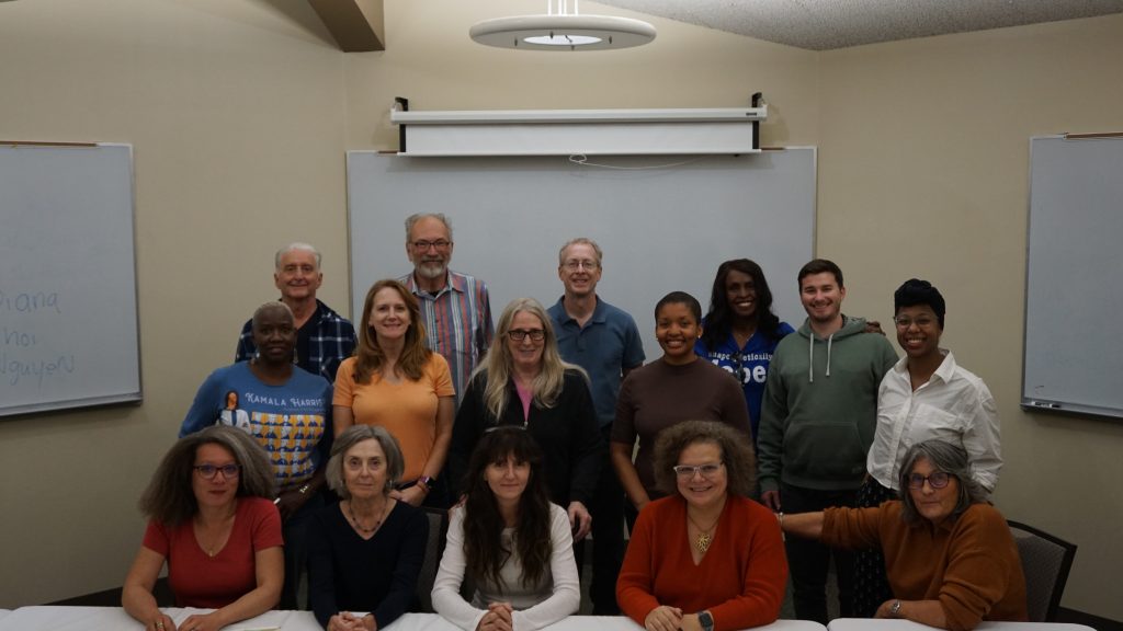 Group photo of workshop participants posed in a classroom or meeting room with whiteboards behind them. Some people are standing and others are seated at a table in front, smiling toward the camera in a casual, community-focused setting.