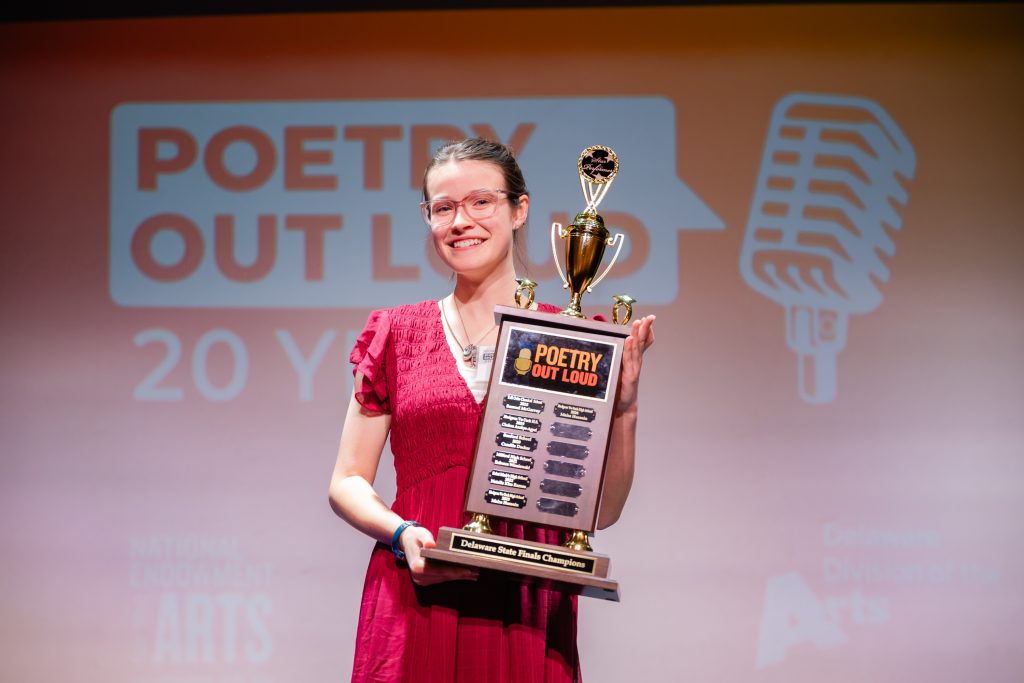 A smiling student in a red dress holds a large “Poetry Out Loud” championship trophy onstage, with the “Poetry Out Loud” logo and a microphone graphic projected on the screen behind them.