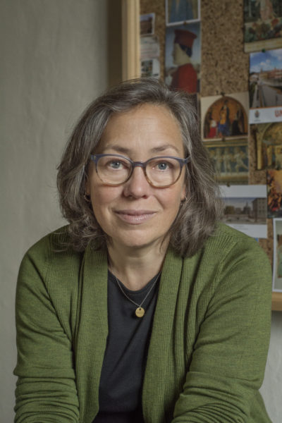 Portrait of a woman with shoulder-length gray hair and glasses, wearing a green cardigan over a dark top and a small pendant necklace. She faces the camera with a gentle, thoughtful expression, seated in front of a bulletin board covered with postcards and small images.