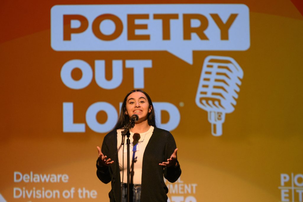 A student stands at a microphone onstage, gesturing while reciting, with the large “Poetry Out Loud” logo projected on an orange screen behind them.