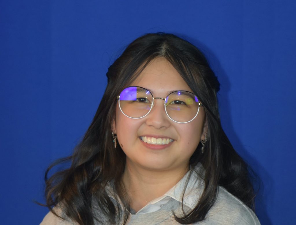 A smiling student with long dark hair and round glasses poses for a headshot against a solid blue background, wearing a light gray sweatshirt over a collared shirt.