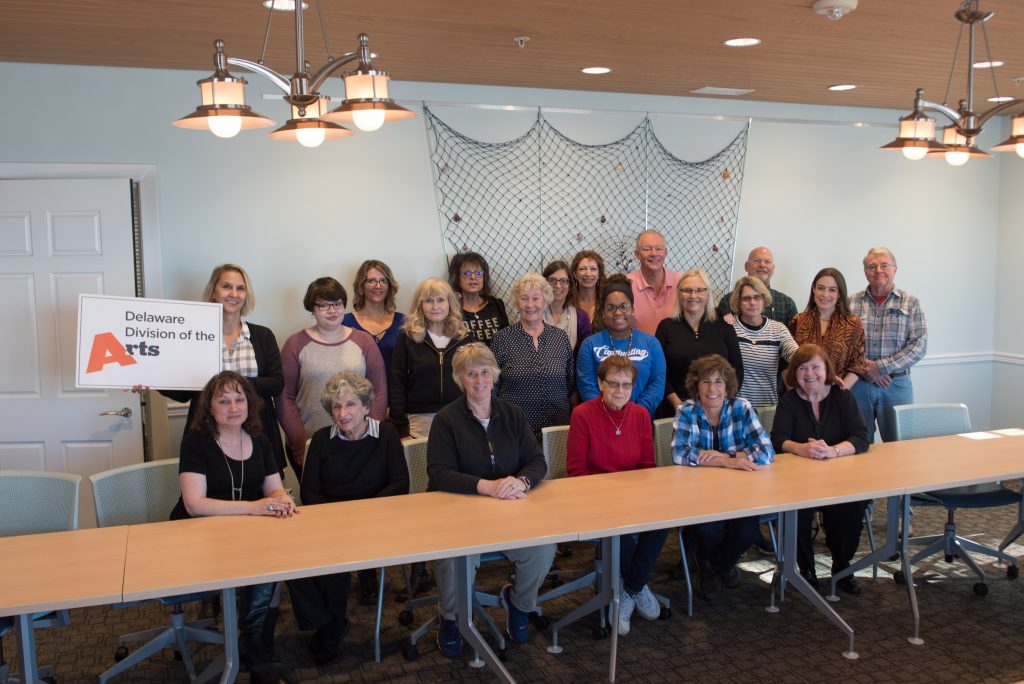 Group photo of Delaware Division of the Arts participants gathered around a long conference table in a meeting room. One person holds a Delaware Division of the Arts sign, while the group smiles toward the camera beneath hanging light fixtures in a bright, casual workshop setting.