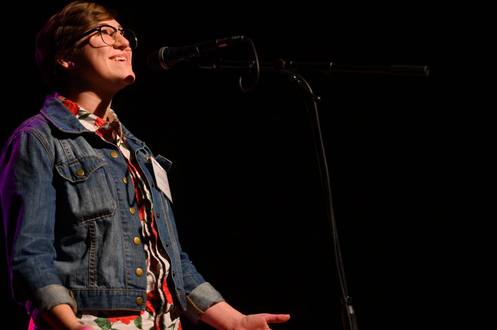 A student performer smiles while speaking into a microphone onstage, wearing glasses and a denim jacket over a patterned shirt against a dark background.