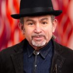 Headshot of a man in a black fedora and jacket against a red-and-orange background.