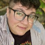 Close-up portrait of a person wearing large black-framed glasses and an eyebrow piercing, with short curly hair and shaved sides, photographed outdoors with greenery softly blurred behind them.