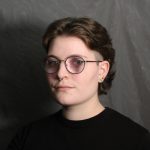 Studio headshot of a person with short brown hair wearing round glasses and a septum ring, dressed in a black top against a gray backdrop.
