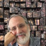 Smiling man with a gray beard and round glasses in front of an abstract patterned wall.