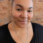 Headshot of a person with closely cropped hair and a septum ring, wearing a black top and a long necklace with a round pendant, standing in front of a softly blurred brick wall.