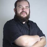 Studio portrait of a bearded person with a septum ring wearing a black button-down shirt, arms crossed, against a light gray background.