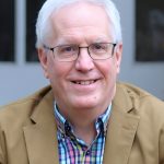 Headshot of a smiling man with white hair and glasses, wearing a tan blazer over a multicolored plaid shirt, with a softly blurred background.