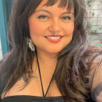 Close-up portrait of a smiling woman with long dark hair and bangs, wearing a black top and a septum ring, photographed outdoors near a brick wall with climbing greenery.