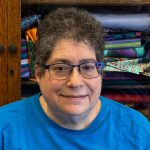 Headshot of a person with short curly hair wearing blue-framed glasses and a bright blue shirt, photographed indoors in front of shelves of colorful folded fabric.