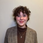 Headshot photo of a person in a brown shirt, tan blazer, and curly brown hair.