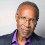 Headshot of a man with short gray hair and glasses, wearing a navy blazer over a purple shirt, smiling gently against a light gray background.