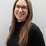 Portrait of a smiling woman with long light-brown hair wearing glasses and a black top, photographed against a light gray background.