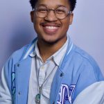Portrait of a smiling man with short curly hair and round glasses wearing a light blue varsity jacket over a white shirt, against a light blue background.