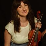 Portrait of a smiling woman with long brown hair holding a violin and bow, wearing a light top and blue skirt against a dark background.