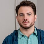 Alt text (recommended): Headshot of a man with short dark hair and a trimmed beard, wearing a teal button-down shirt and a blue hoodie, looking at the camera against a neutral indoor background.
