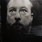 Black-and-white close-up portrait of a bald person with a full beard, gazing slightly upward against a dark, textured background.