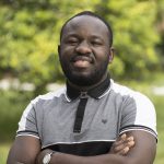 Portrait of a smiling man with a short beard standing outdoors with his arms crossed, wearing a gray-and-black polo shirt, with a softly blurred green background.
