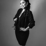 Black-and-white studio portrait of a smiling woman with a long ponytail, wearing a dark zip-front jacket over a fitted dress, posing with one hand on her hip against a plain gray backdrop.