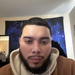 Close-up selfie of a person with short dark hair and a full beard, wearing a tan hoodie with a cream-colored hood, indoors with a galaxy-pattern tapestry on the wall behind them.