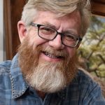 Close-up headshot of a smiling man with blond hair, black rectangular glasses, and a full reddish-blond beard, wearing a blue plaid shirt indoors.