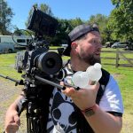 Man outdoors beside a professional film camera, holding two water bottles.