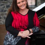 Portrait of a smiling woman leaning on a grand piano, wearing a red-and-black patterned top, photographed indoors with a stone wall and window softly blurred in the background.