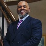 Portrait of a smiling man in a dark suit with a pink-and-blue striped tie, standing indoors near a staircase. Alt text (short):