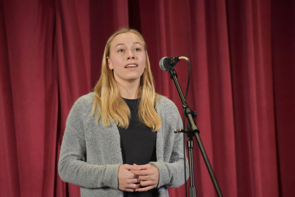 A student with long blonde hair stands onstage in front of red curtains, speaking into a microphone on a stand while wearing a gray cardigan over a dark top.