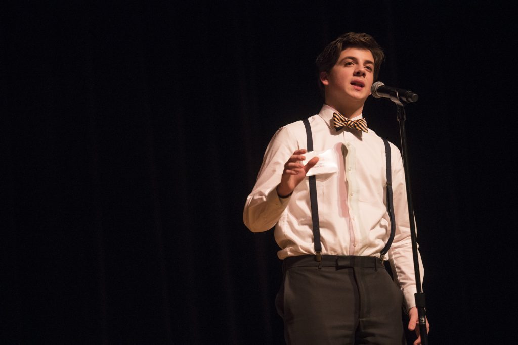 A student performer stands at a microphone on a dark stage, wearing a white dress shirt with suspenders and a striped bow tie, gesturing while reciting.