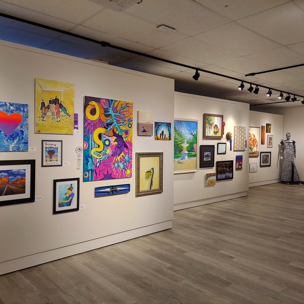 Wide view of a gallery exhibition featuring a diverse collection of framed paintings, photographs, and mixed-media works displayed across white walls under track lighting. The open room includes colorful abstract and figurative pieces in many sizes, with gray wood-look flooring and a full-length sculptural garment or figure visible at the far right.
