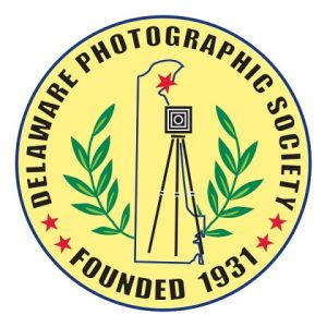 Circular logo for the Delaware Photographic Society with the words “DELAWARE PHOTOGRAPHIC SOCIETY” around the top and “FOUNDED 1931” around the bottom; inside is the outline of Delaware with a camera on a tripod, laurel branches on both sides, and small red stars.