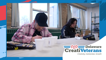 Wide promotional banner framed in blue showing a CreatiVeterans art workshop. In the foreground, a person in a black cap and red plaid shirt focuses on drawing or writing at a table with art supplies and cups. In the background, another participant works quietly at the same table. Bottom right includes the “Delaware CreatiVeterans” logo with the tagline “Creativity. Connection. Growth.”