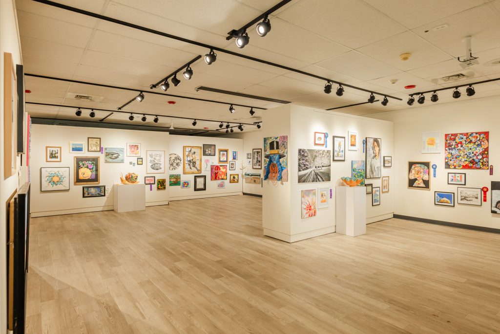 Wide view of a spacious gallery exhibition with framed artworks and a few sculptural pieces displayed on white walls and pedestals throughout the room. Paintings, drawings, and mixed-media works in many styles fill the space under track lighting, with open floor area and light wood flooring creating a clean, welcoming setting.