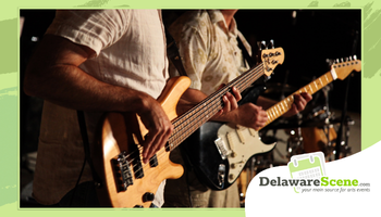 Close-up of musicians performing live, focusing on hands playing an electric bass guitar in the foreground and an electric guitar in the background under warm stage lighting. A green graphic border frames the image, with the DelawareScene.com logo and tagline in the lower right corner.