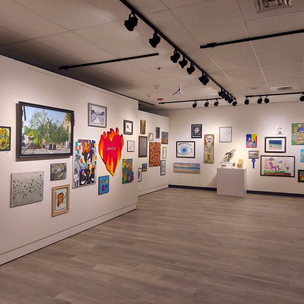 Wide view of a gallery exhibition with paintings, drawings, and mixed-media works displayed across white walls under black track lighting. The room includes framed artworks of many sizes, a bright heart-shaped piece, and a small sculpture on a pedestal, all set within an open space with gray wood-look flooring.