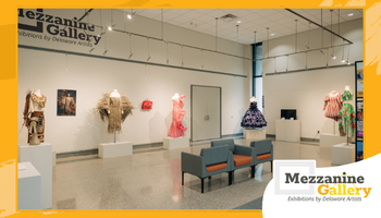 Wide view of a bright, modern gallery space with a polished floor, white walls, and track lighting. Artworks—primarily colorful wearable/fiber pieces and garments—are displayed on pedestals and mannequins along the left and back walls, with additional pieces on the right. Two teal lounge chairs sit in the center. An orange graphic border frames the image, and a “Mezzanine Gallery: Exhibitions by Delaware Artists” logo appears in the lower right.