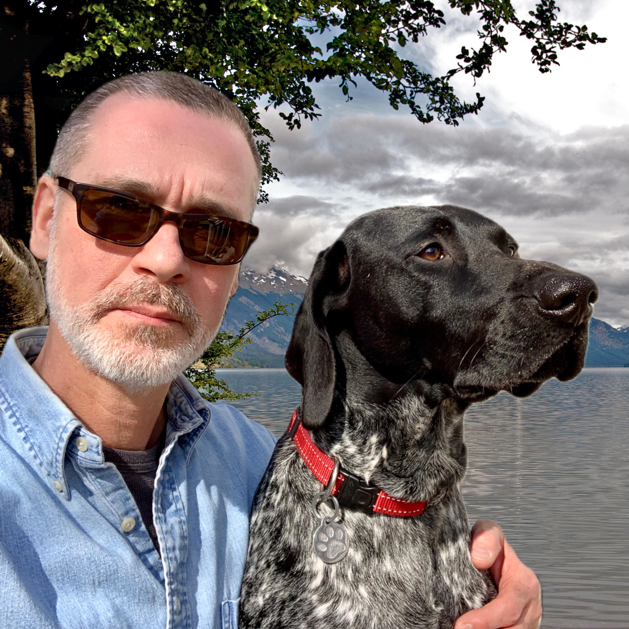 Outdoor selfie of a man wearing sunglasses and a light blue button-down shirt holding a black-and-white speckled dog with a red collar and tag.