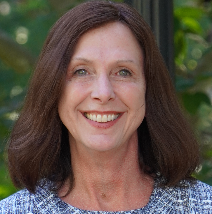 Headshot of a smiling woman with shoulder-length brown hair wearing a patterned top, photographed outdoors with greenery in the background.