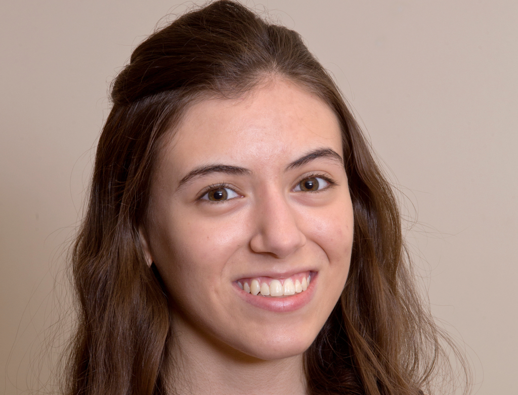 Close-up headshot of a smiling student with long brown hair against a light, neutral background.