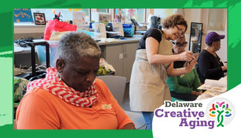 Wide promotional banner framed in green showing a Creative Aging workshop in a classroom. In the foreground, an older man in an orange shirt with a red-and-white scarf looks down while seated at a table. In the background, a woman stands and helps an older participant with a hands-on activity, while other older adults work at tables near computers. Bottom right includes the “Delaware Creative Aging” logo with a colorful tree/people icon.