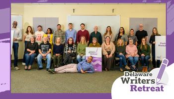 Wide promotional banner framed in purple showing a large group photo indoors. Around two dozen adults pose in rows—some seated on chairs, others standing behind—while one person lies playfully on the floor in front. Several people hold small signs. In the lower-right corner, a logo reads “Delaware Writers Retreat” with a pencil icon.